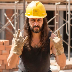 Fotografia de médio plano do Engenheiro Civil Diego Badaró em um canteiro de obras. Ele tem cabelo longo e barba, veste um capacete de segurança amarelo e uma regata preta desgastada. Ele está fazendo o gesto de "chifres do rock" com as duas mãos, que estão calçadas com luvas de couro sujas e desgastadas. Sua expressão é séria e direta para a câmera. O fundo mostra tijolos empilhados e andaimes de metal sob luz solar forte.